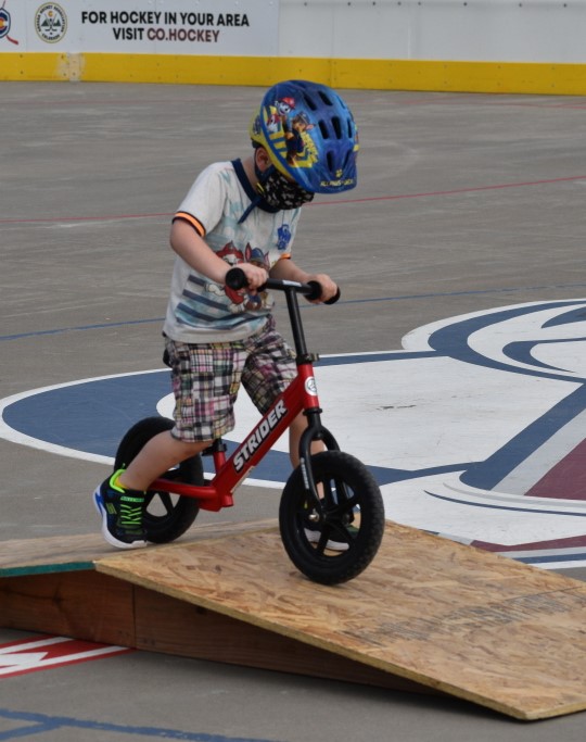 child wearing a helmet, riding a strider bike up a small wooden ramp.